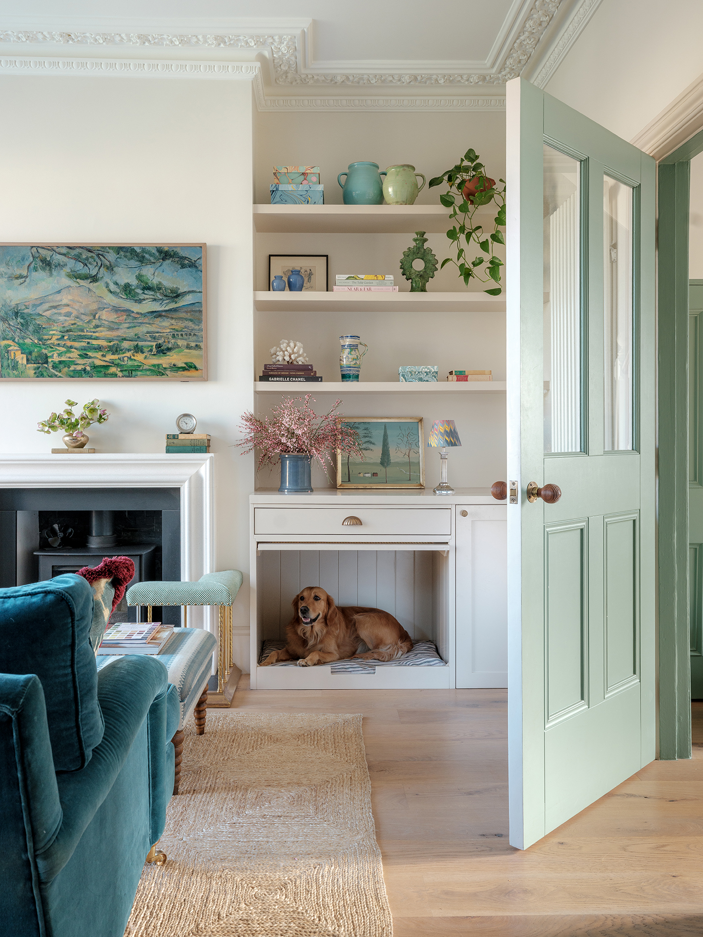 A living room with pale pink walls and light wood floors. In the foreground the corner of a green-blue velvet sofa can be seen. Built-in shelves display vases and trinkets and beneath, a dog lays in a dog nook built into the cabinetry 