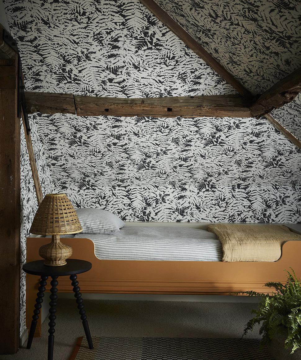 a bedroom with white and black botanical wallpaper, wooden beams, an orange painted bed frame with white stripe bedding and a small bedside table with a rattan lamp