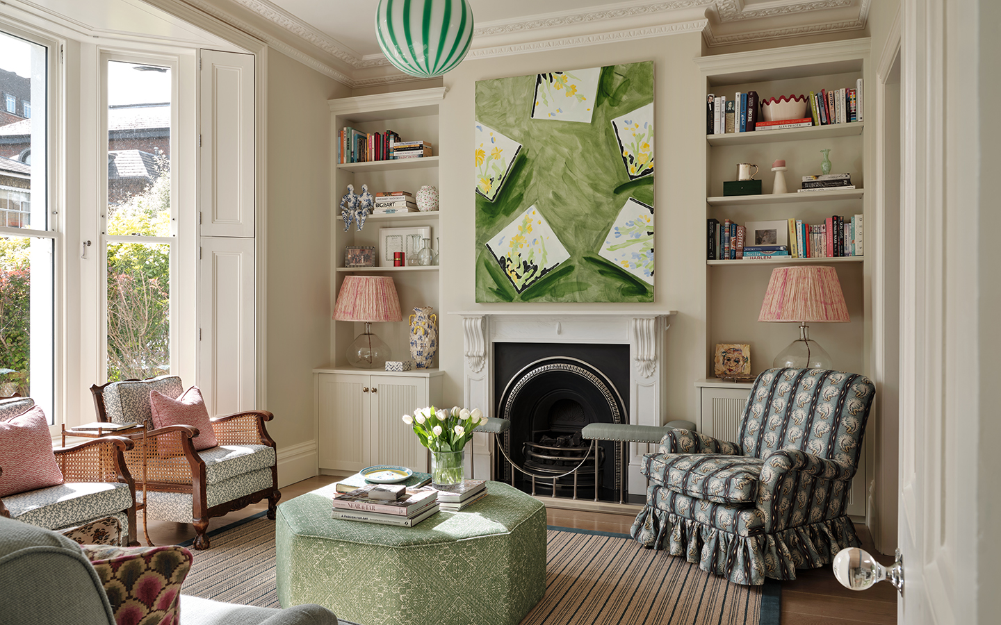 A living room with an octagonal green ottoman in the centre surrounded by a patterned frilled armchair, two cane armchairs and a blue sofa seen in the bottom corner, all arranged on a striped rug. On the back wall is a fireplace and large statement artwork