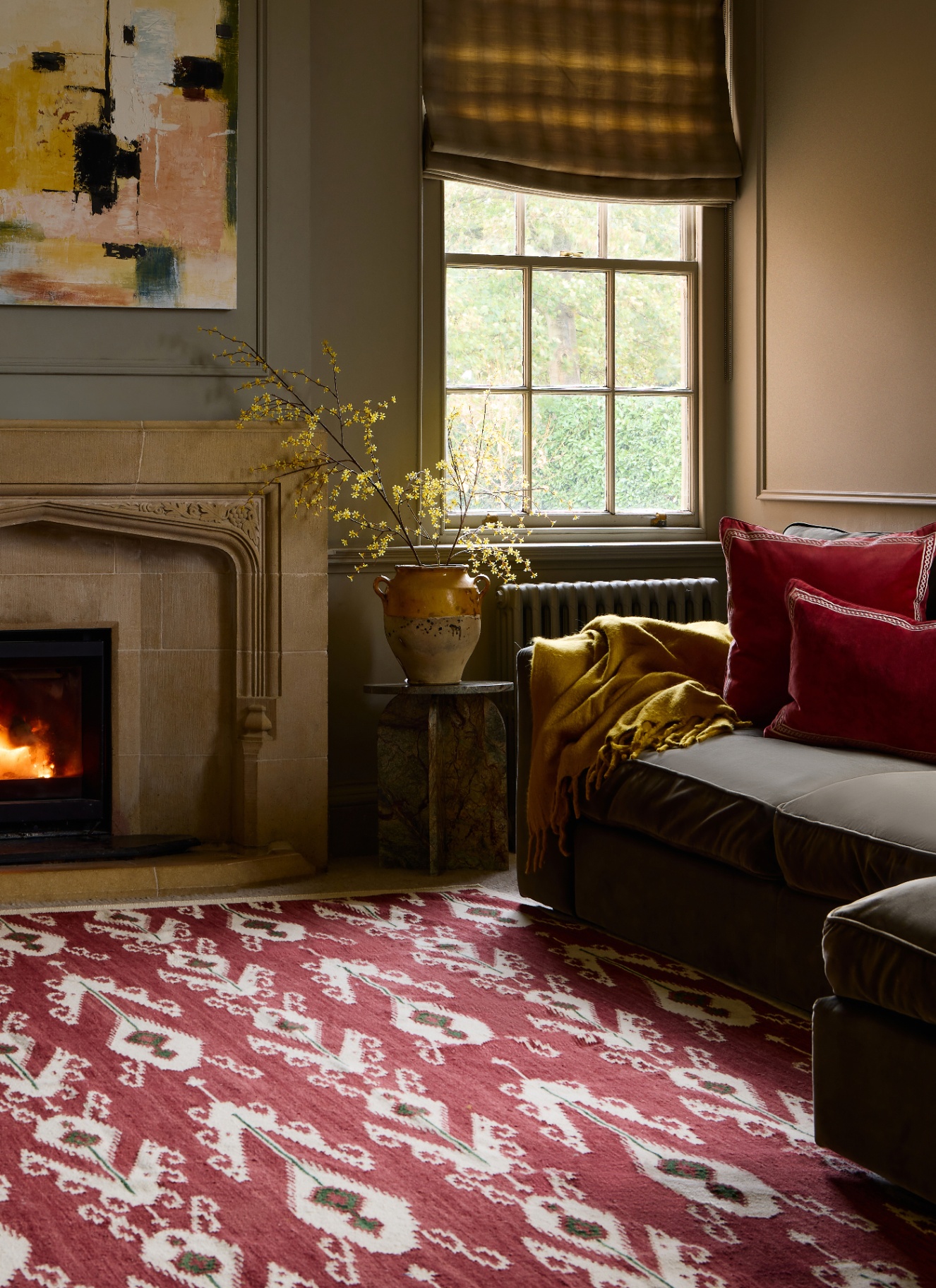 A warm, cosy living room with a red patterned rug on the floor, a brown sofa with red cushions and a mustard yellow throw, a carved stone fireplace with a glowing fire, and a sash window
