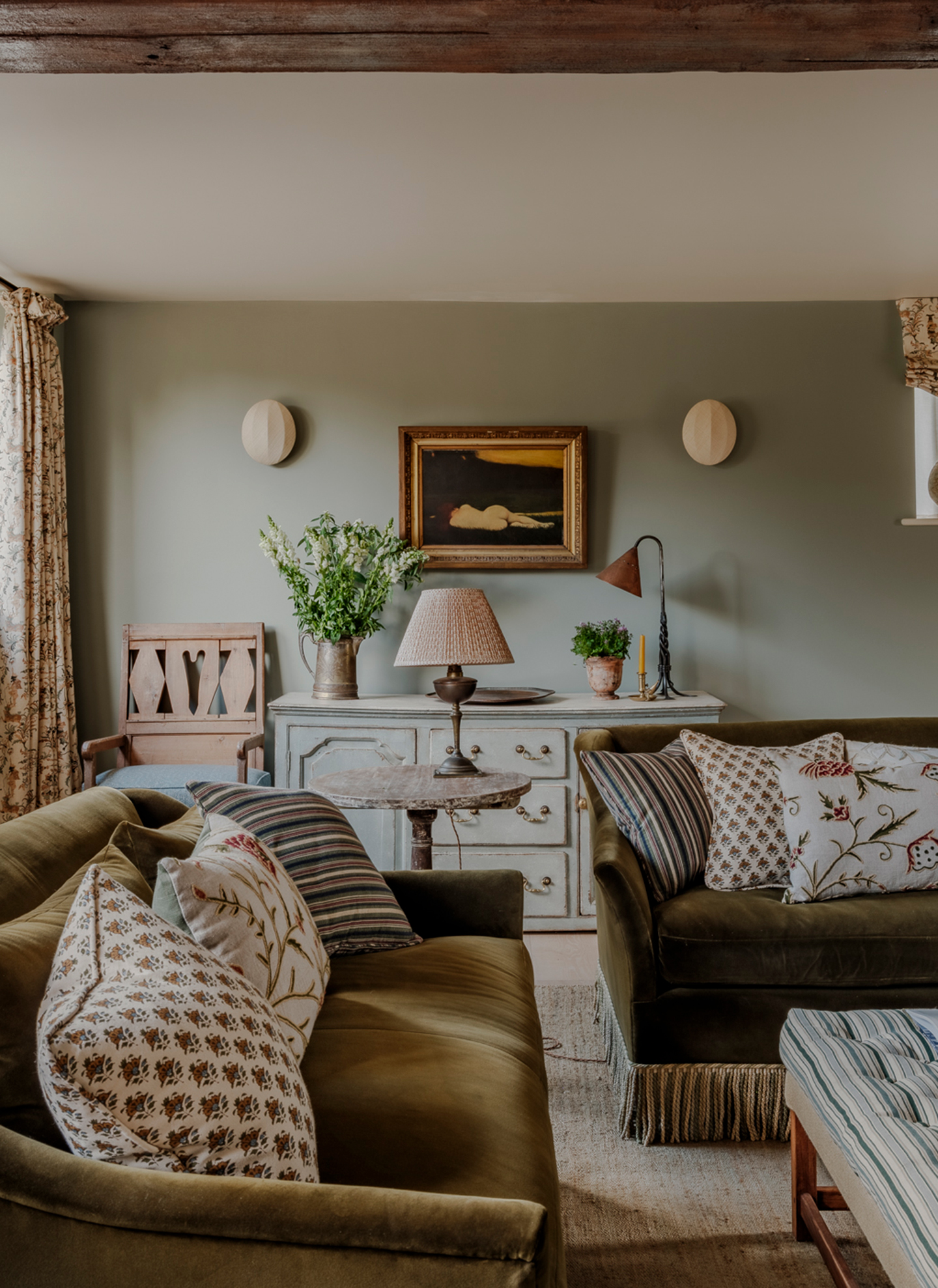 a living room with pale green walls, two olive green fringed sofas and a pale green sideboard