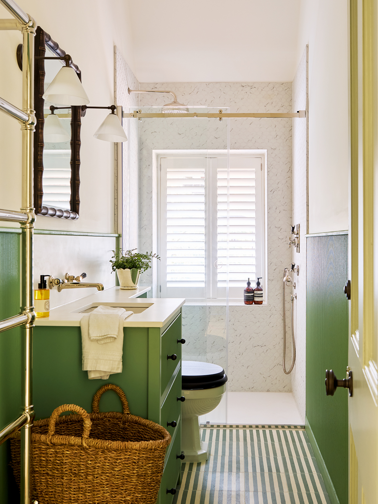 A shower room with a marble enclosed shower at the far end, and a toilet, built-in basin and large wicker basket in the foreground. The floor tiles are green and white striped, and the walls are green on the lower half and white on the upper half