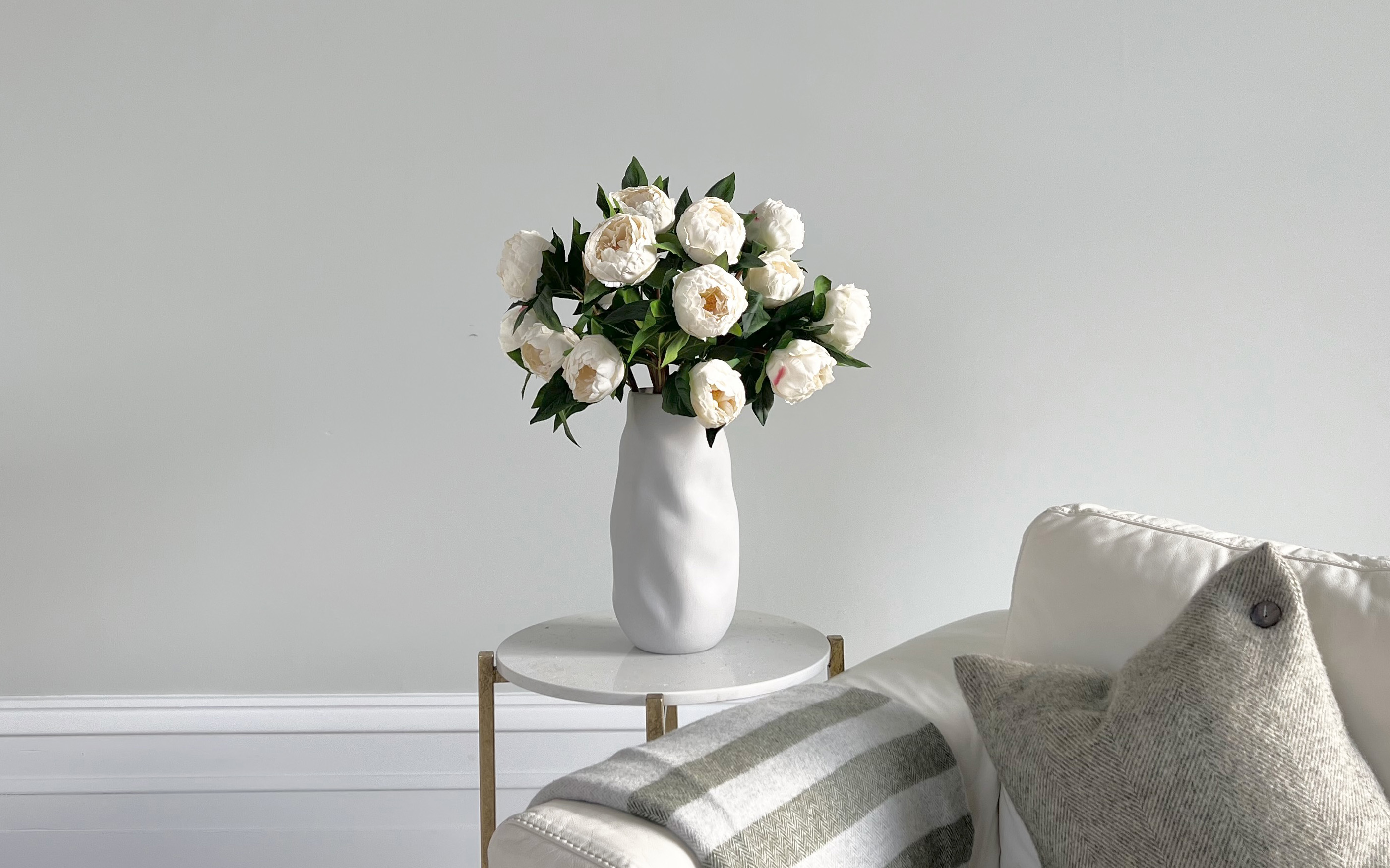 a bunhch of white faux peonies in a white vase on a white side table next to a white sofa with a stripe throw over the arm