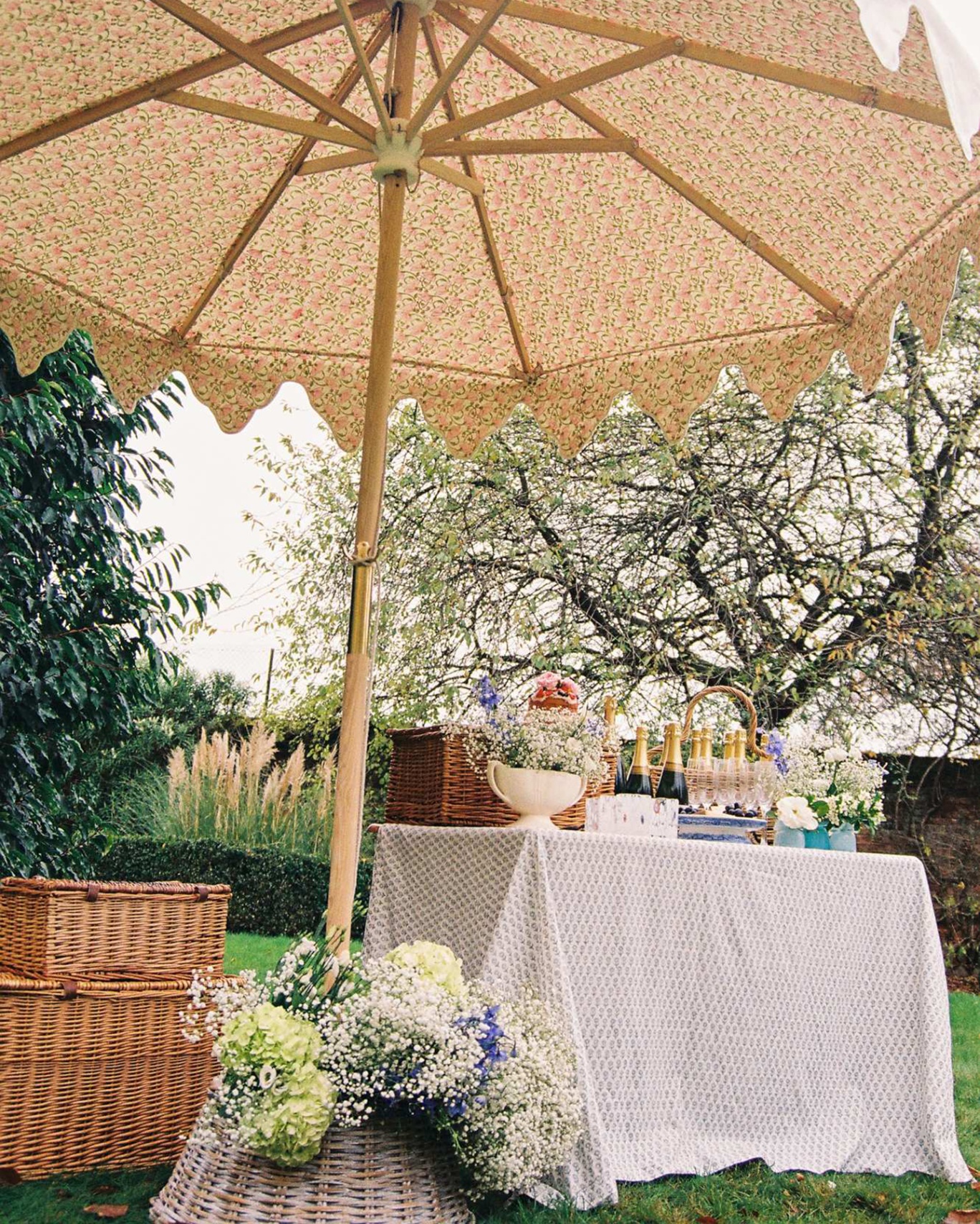 an outdoor table with a table cloth and bottles of champagne and champagne flutes under a patterned parasol