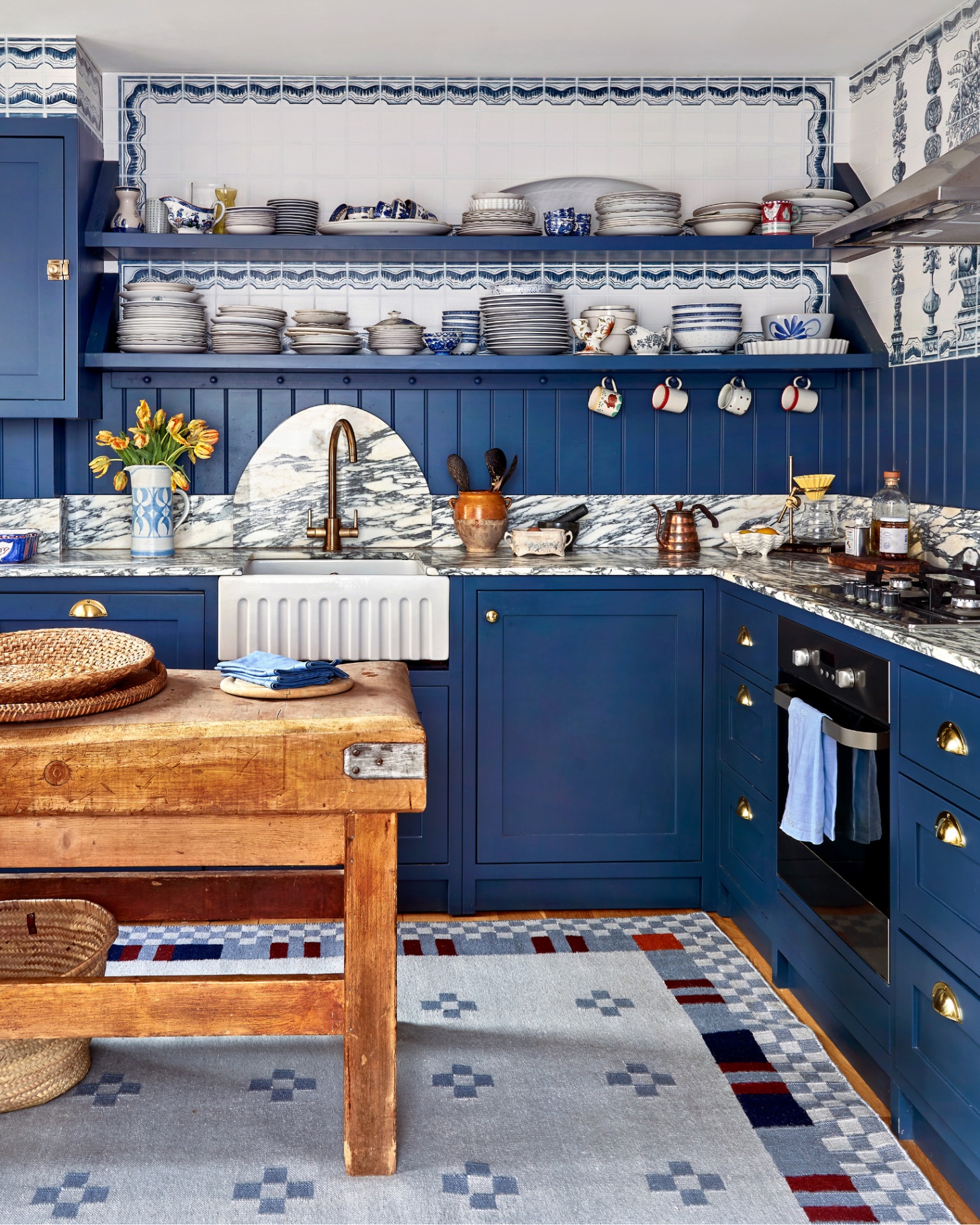 a kitchen with a large blue patterned jute rug under a reclaimed wood table, blue kitchen cabinets with white marble tops, blue and white patterned tile walls and open shelves with stacks of plates