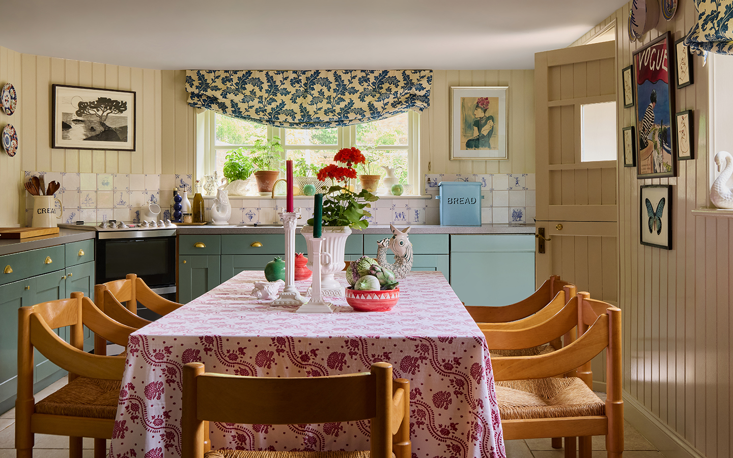 A kitchen with a large table covered with a floral cloth and dining chairs in the foreground. In the background, lower cabinets line the tongue-and-groove walls, a tiled backsplash above. A botanical print blind dresses a large window 