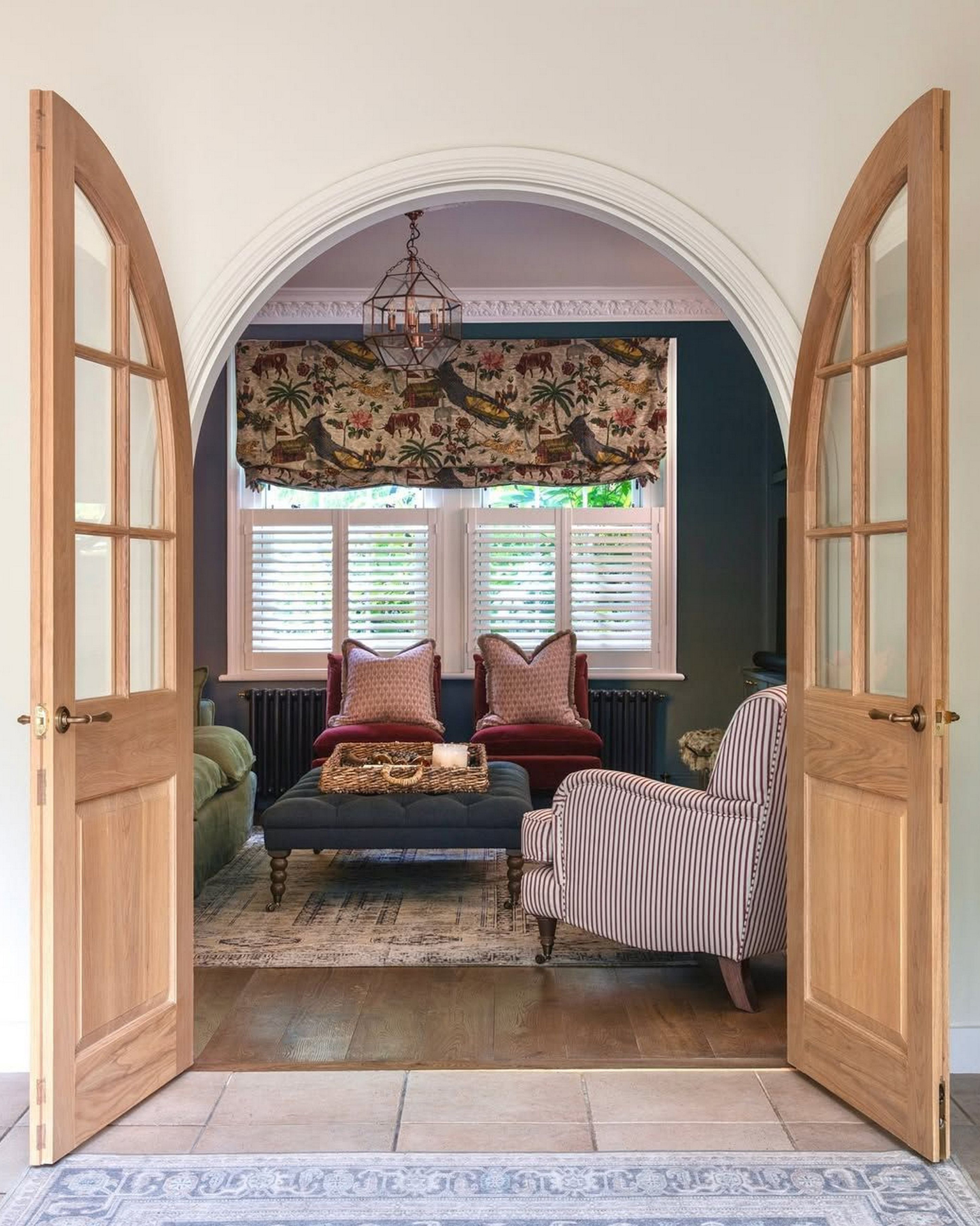 A living room seen through open arch doors, a wooden floor with a persian rug, an upholstered coffee table, two red velvet chairs with large cushions and a back window with shutters and a cream jungle patterned blind