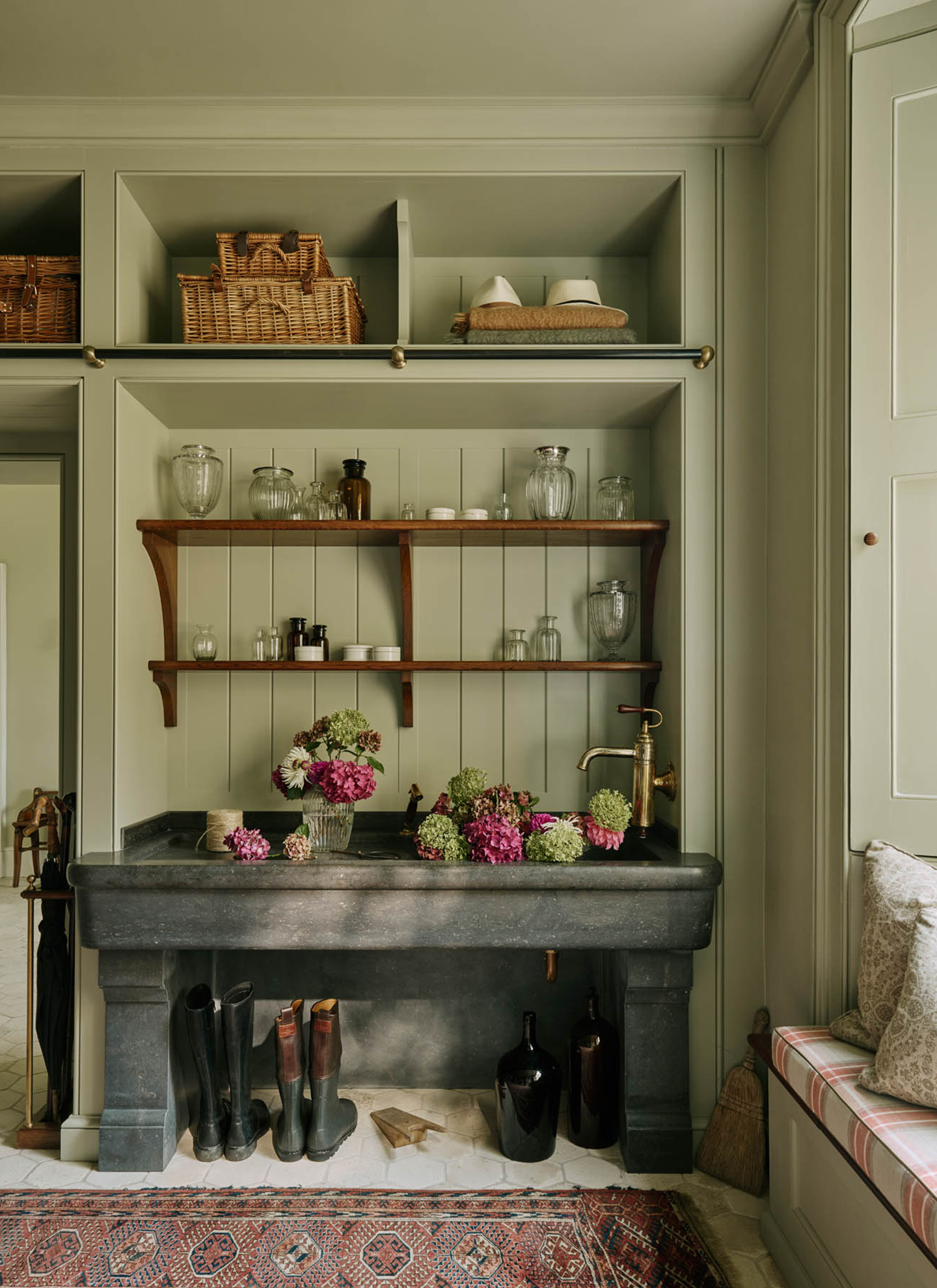 a bootroom with sage green paneled walls, a large dark grey stone sink with flowers, open wooden shelving and wellington boots under the sink