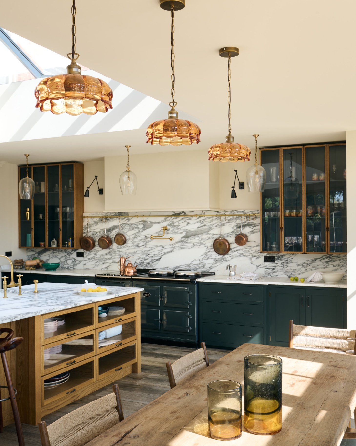 a kitchen with 3 amber glass pendant lights hanging between a wooden dining table and chairs and a wooden kitchen island with a white and grey marble top