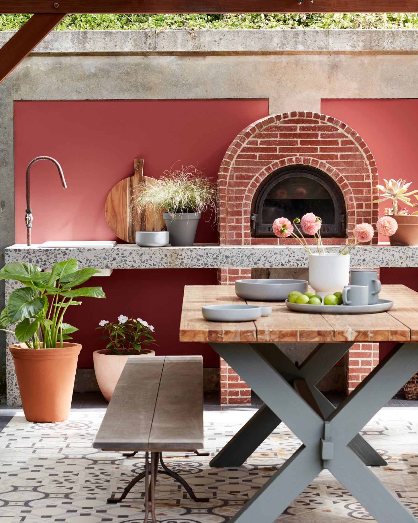 an outdoor kitchen with walls painted in tuscan red, an integrated pizza oven with a brick surround and a wooden outdoor dining table with grey legs and a dining bench