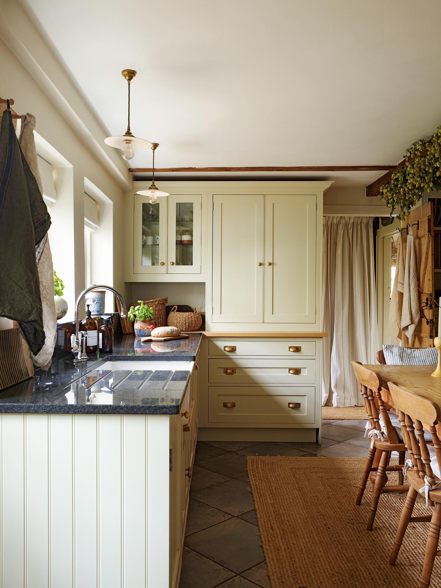 a neutral country kitchen, with cabinets running along the left and a tall built-in cabinet at the back. Two small pleat ceramic lights hang above the kitchen worktops. On the right the corner of a wooden dining table and chairs sits on a jute rug