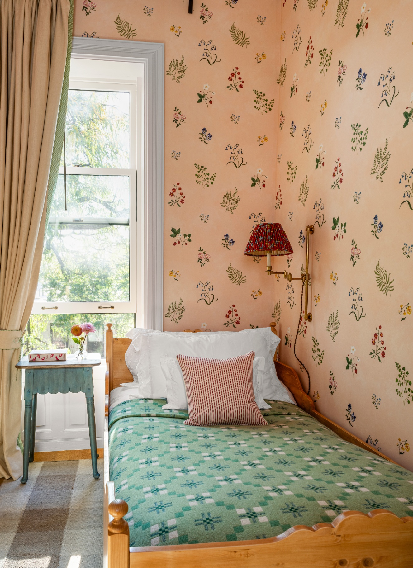 A small single bedroom with floral wallpaper, featuring an articulating brass wall light with a red patterned pleated shade mounted near the headboard