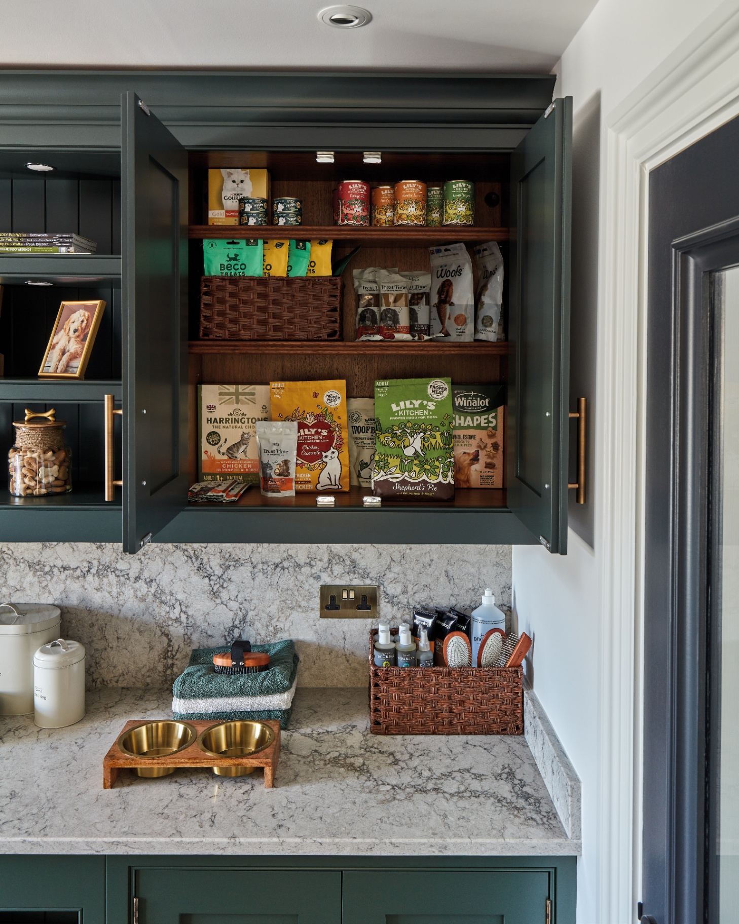 a kitchen cupboard pantry painted in dark green with the cupboard filled with dog food and the white masrble countertop with a gold dog bowl and basket full of dog grooming products