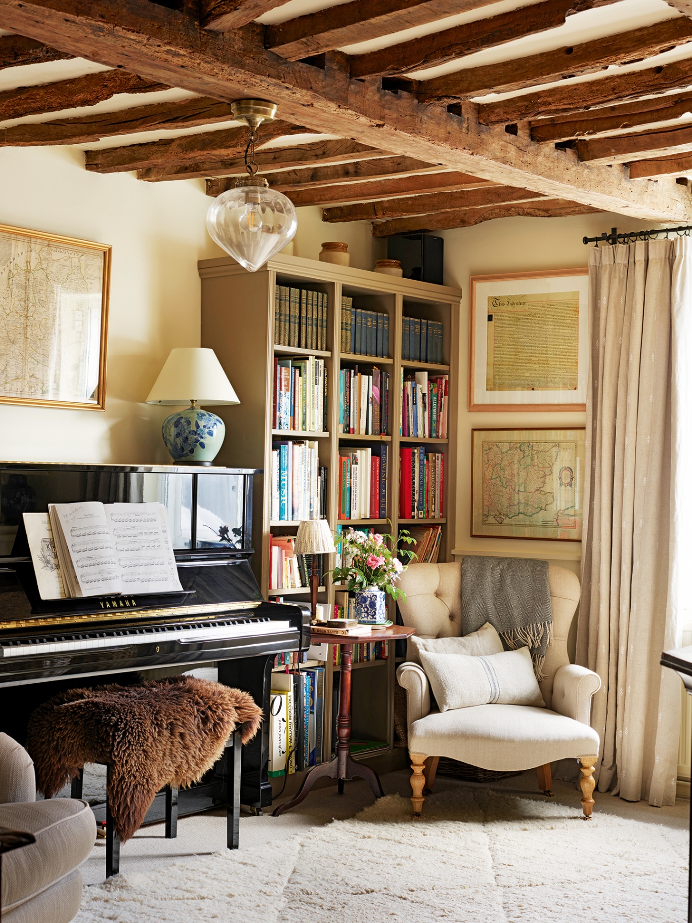 A corner of a living room with wooden ceiling beams and a tufted white carpet. A bookshelf sits in the corner with an armchair and side table in front and a piano topped with a table lamp to the side. A glass pendant hangs above