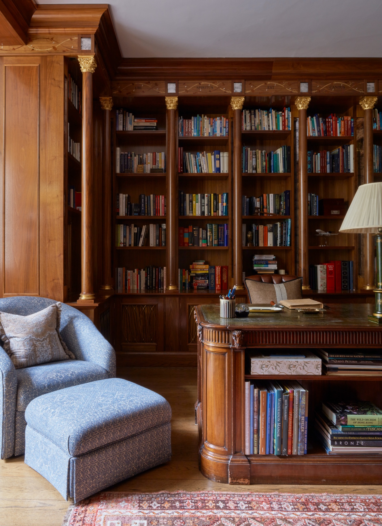 a study with a built in large bookcase in a dark wood with column details, a large dark wooden antique style desk with a blue patterned upholstered chair next to it and a persian rug