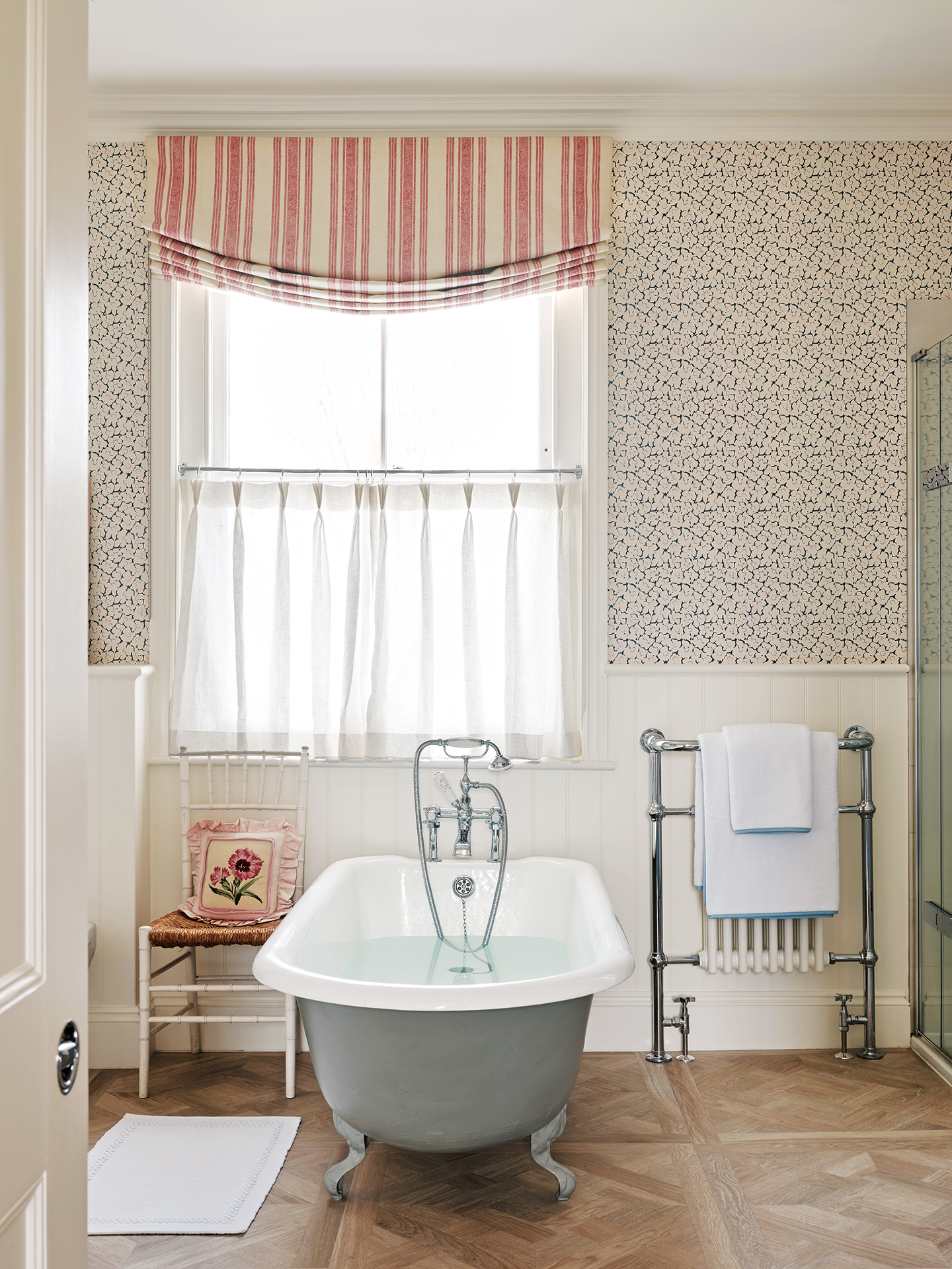 A bathroom with a freestanding bath in the centre, with an antique white chair with a floral cushion to the left and a towel rail to the right. The walls are papered with a small-scale print on the upper half and white tongue and groove on the lower half