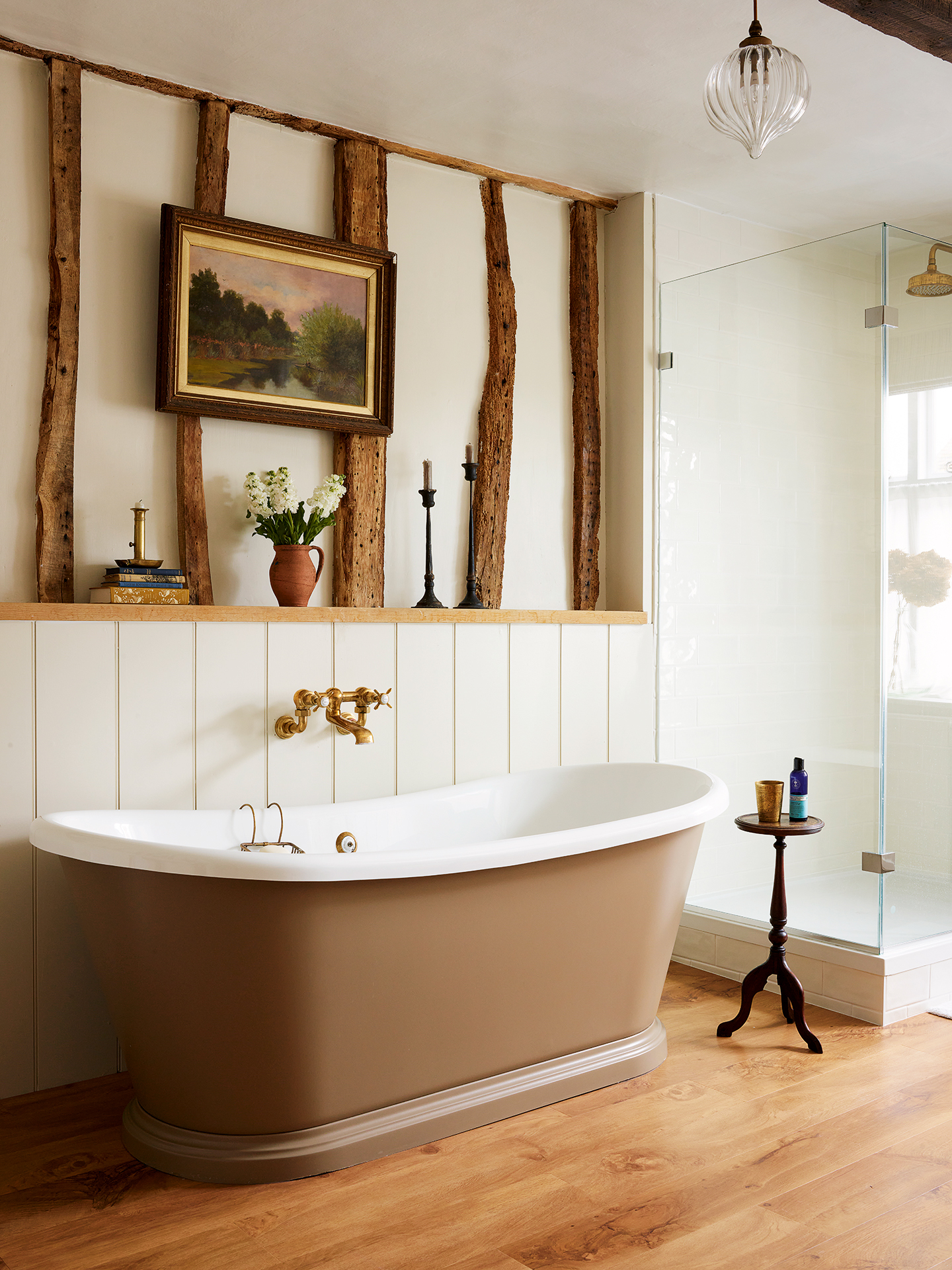 A bathroom with wood floorboards, tongue-and-groove panelling on the lower walls and wooden beams running up the upper half. A brown freestanding bathtub sits below a wall-mounted brass tap. Next to it is a shower and small side table