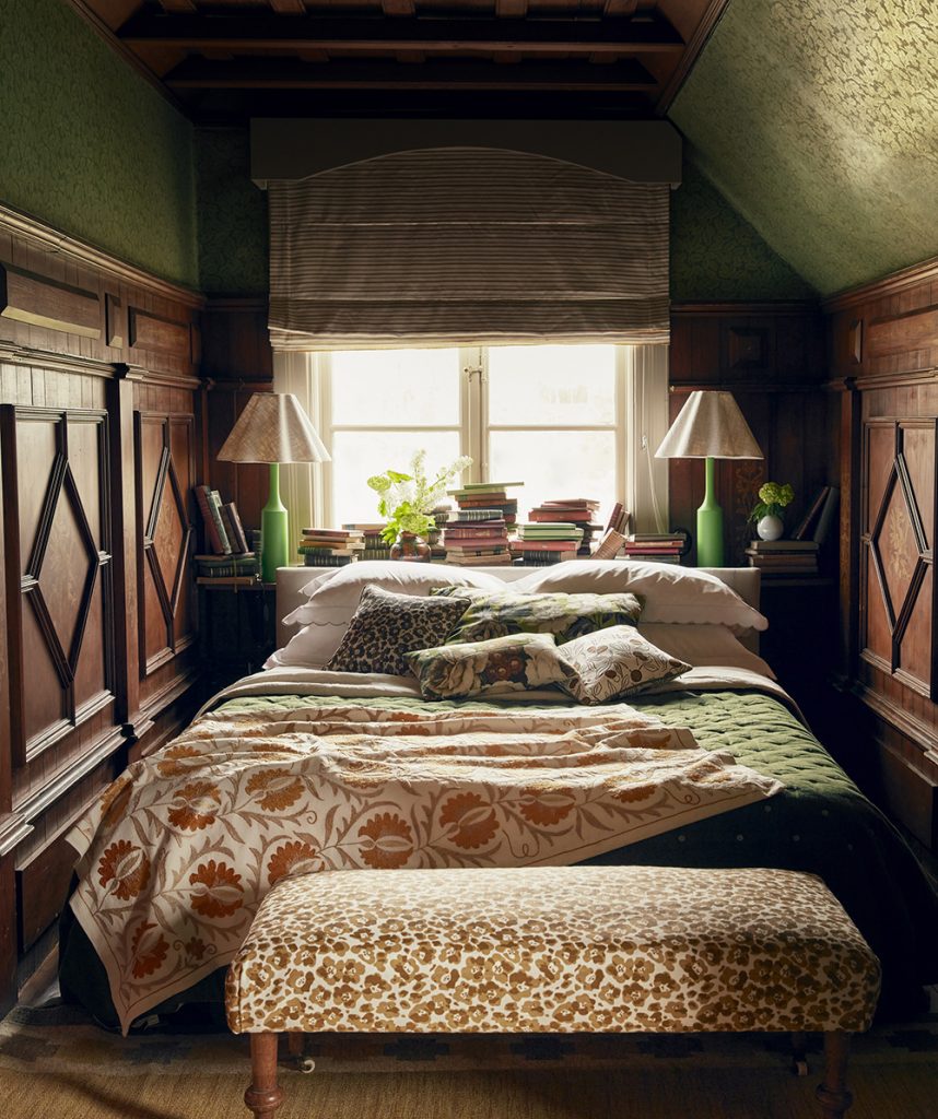 a small bedroom with half dark wood panelled walls and green ditsy floral wallpaper a bed with a green bedspread, an ottoman at the foot of the bed and two large pleated lamps next to the bed