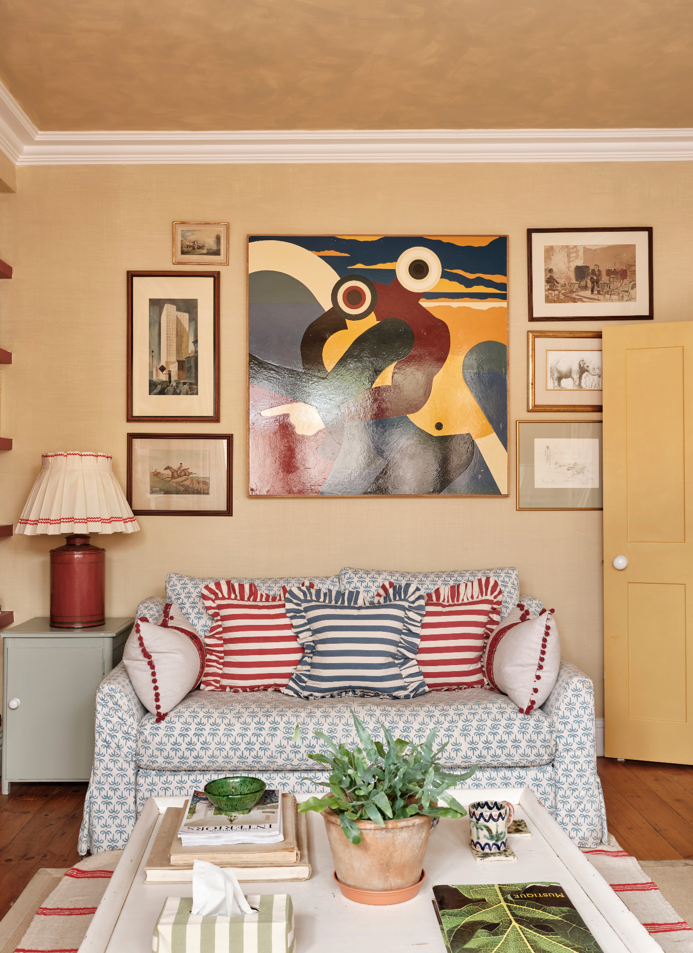 a living room with a white and blue patterned sofa with blue and white striped cushions and red and white striped cushions and beige walls with a large piece of artwork