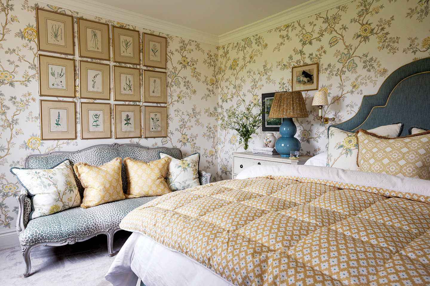 A bedroom covered in floral wallpaper with a double bed to the right, topped with decorative cushions and a yellow eiderdown, and a leopard print bench on the left with a series of twelve botanical artworks on display above