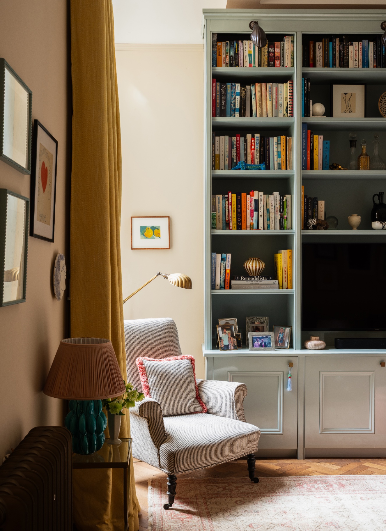 A cosy reading corner featuring a patterned armchair, a tall brass floor lamp, mustard yellow curtains, and large pale-blue built-in bookshelves filled with books.