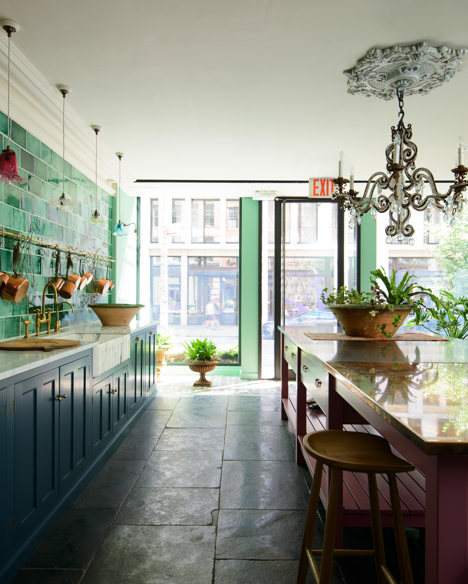 a kitchen with a large grand chandellier hanging above a dark red kitchen island with a bench tucked underneath and a terracotta pot on top, dark blue kitchen cabinetry and green tiled splashback
