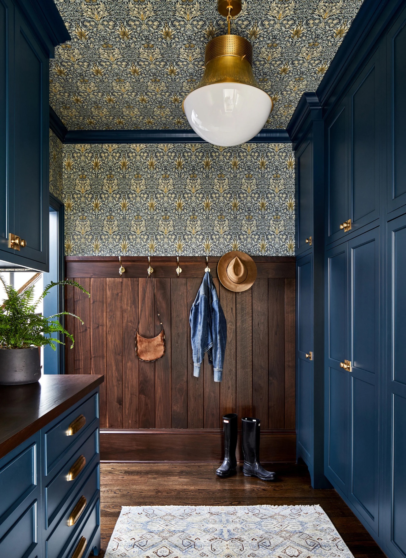 An elegant boot room featuring deep blue fitted cabinets, dark wood wall panelling with brass peg hooks, patterned wallpaper, a brass pendant light, and black wellington boots set on a polished timber floor