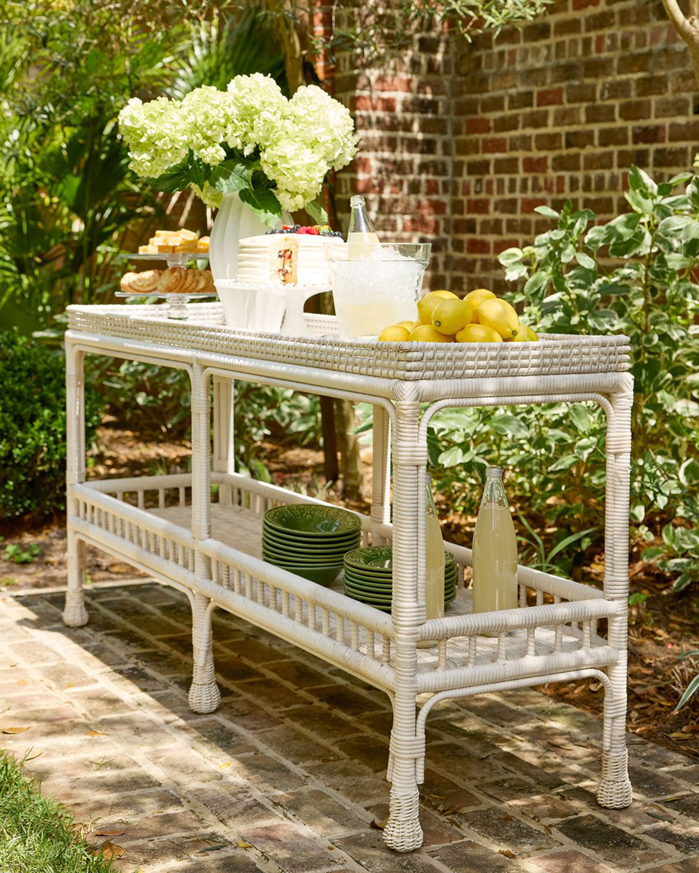 an outdoor white rattan console table filled with drinks, lemons and plates against a red brick wall
