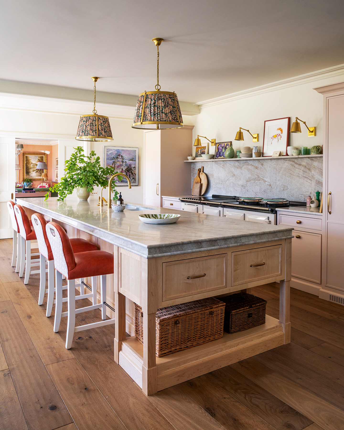 A light kitchen with a large oak and marble island in the foreground, paired with orange leather bar stools, wicker baskets on a lower shelf, and two fabric pendant lights above. In the background, cabinets line the wall with a marble backsplash and Aga