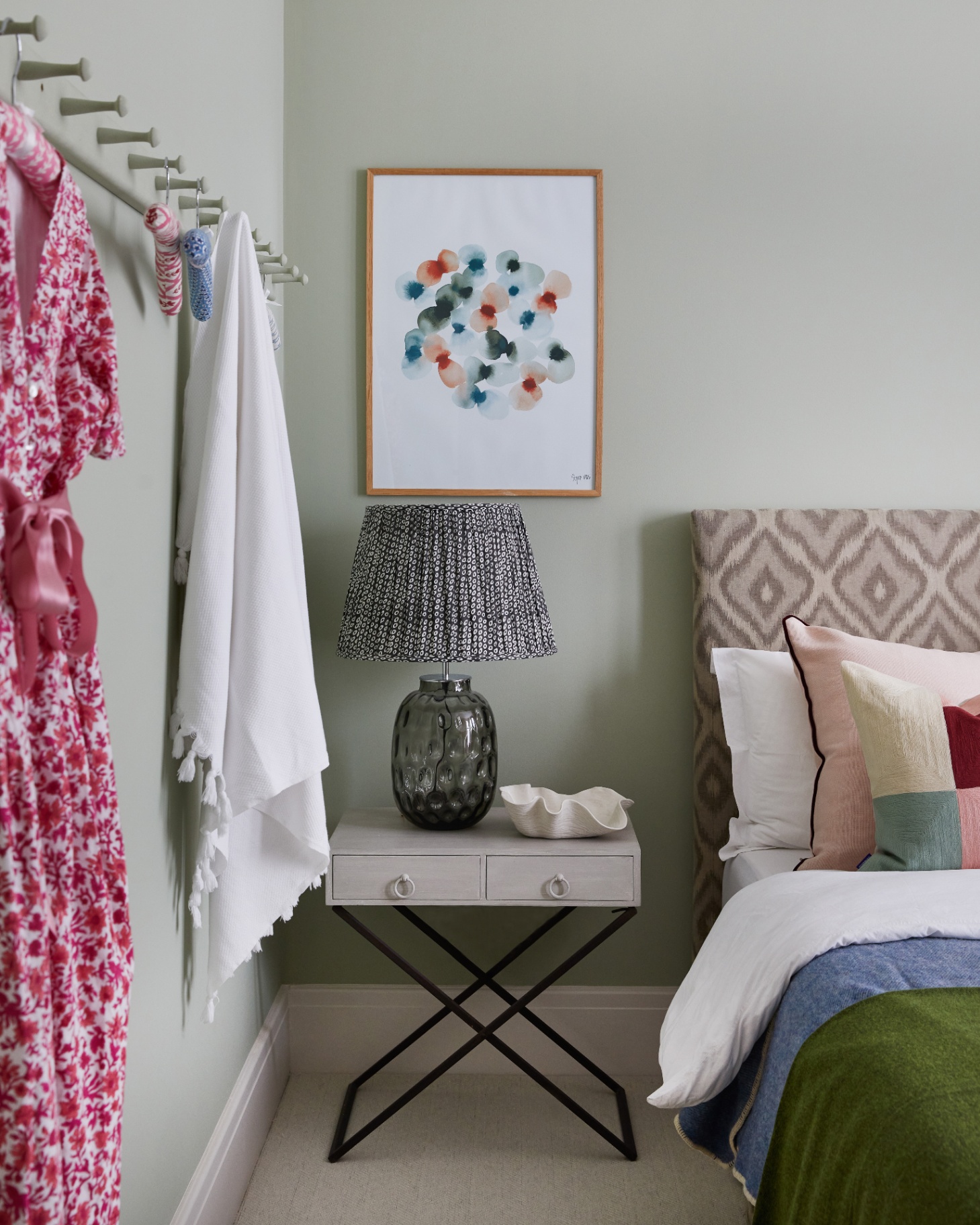 a bedroom with sage green walls, a grey side table with black legs and a large lamp, a bed with a cream patterned headboard and shaker pegs on the wall with a pink dress hanging on it