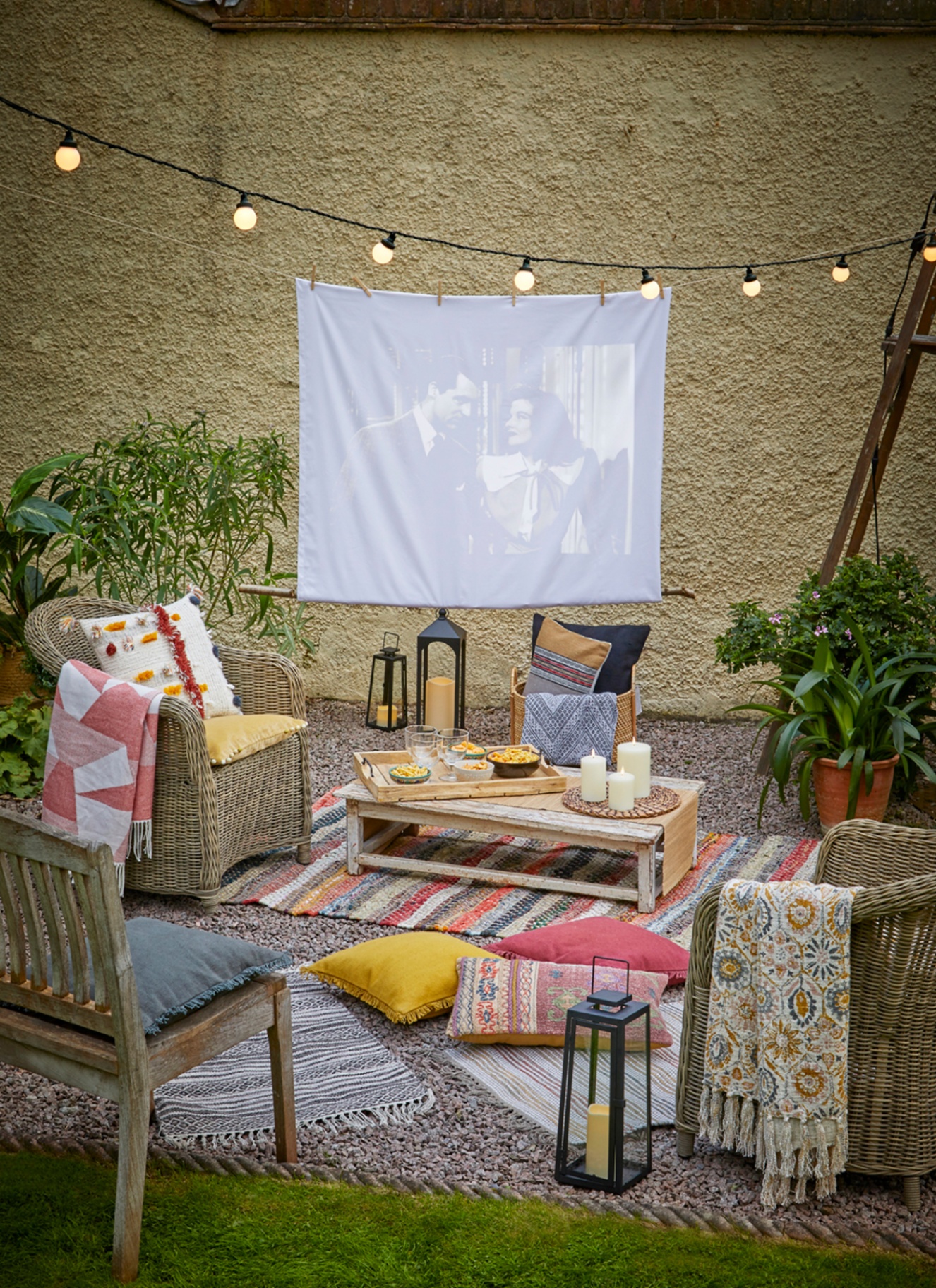 A garden with a white sheet hung up on a line with a movie projected on to it, a striped rug and a coffee table on gravel in front of the screen and wicker and wooden chairs facing the screen