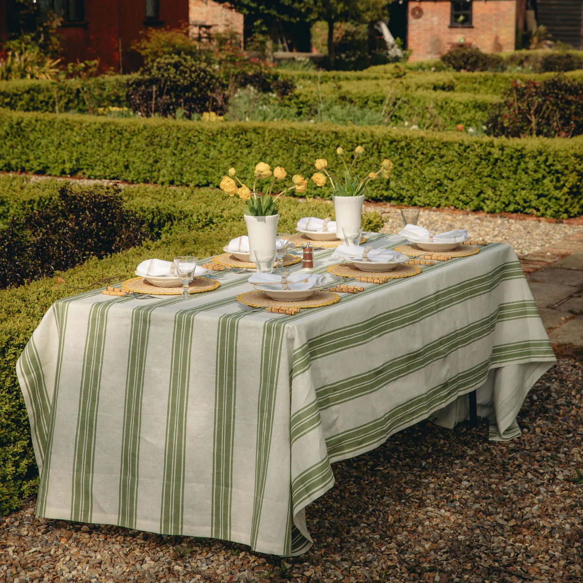 a green striped table cloth on a garden table with a white vase on top with yellow flowers