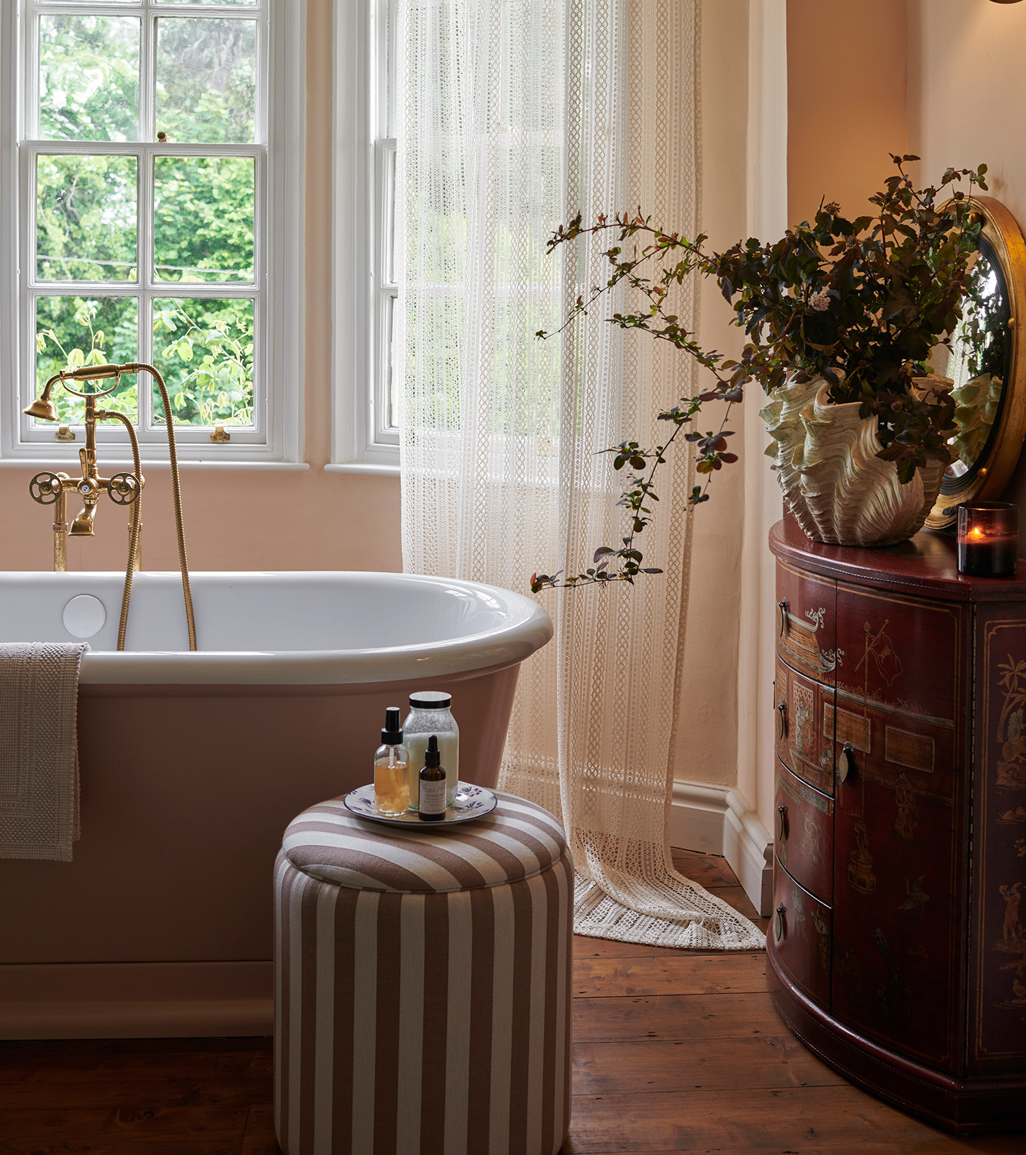 a bathroom with a free standing bath, a large vintage chest of drawers with a shell shaped vase on top with greenery and a white and beige striped ottoman next to the bath