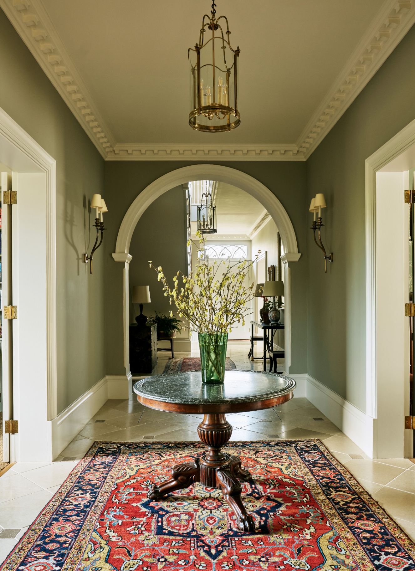 a hallway with sage green walls, a large round table in the centre with a vase of flowers, a lantern light and two sconces on each wall