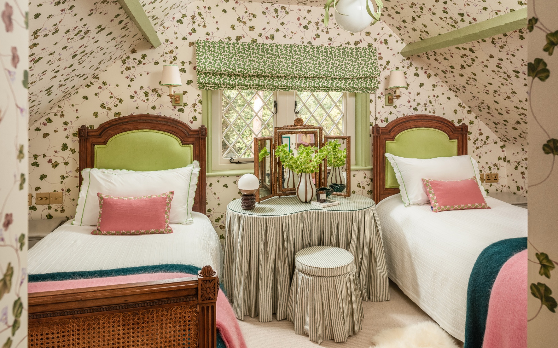 a bedroom with white and green botanical wall paper, two rattan twin beds with white bedding and pink cushions and a large anitque dressing table in front of a window