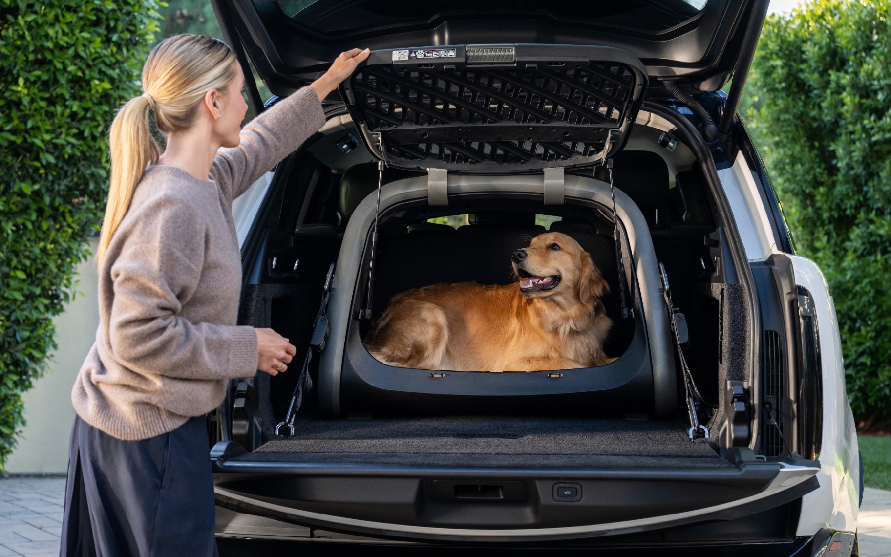 A woman standing at the open tailgate of a modern SUV, holding open a black Tavo Everett dog crate with a Golden Retriever inside