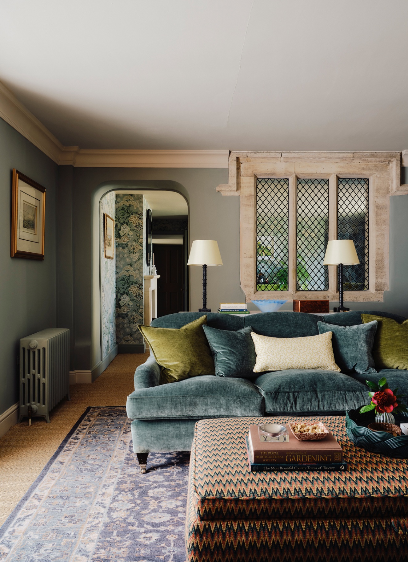 An atmospheric sitting room featuring a deep teal velvet sofa accenting muted sage walls, paired with olive green cushions, a chevron-patterned ottoman coffee table, and an arched doorway leading to floral-wallpapered hallway