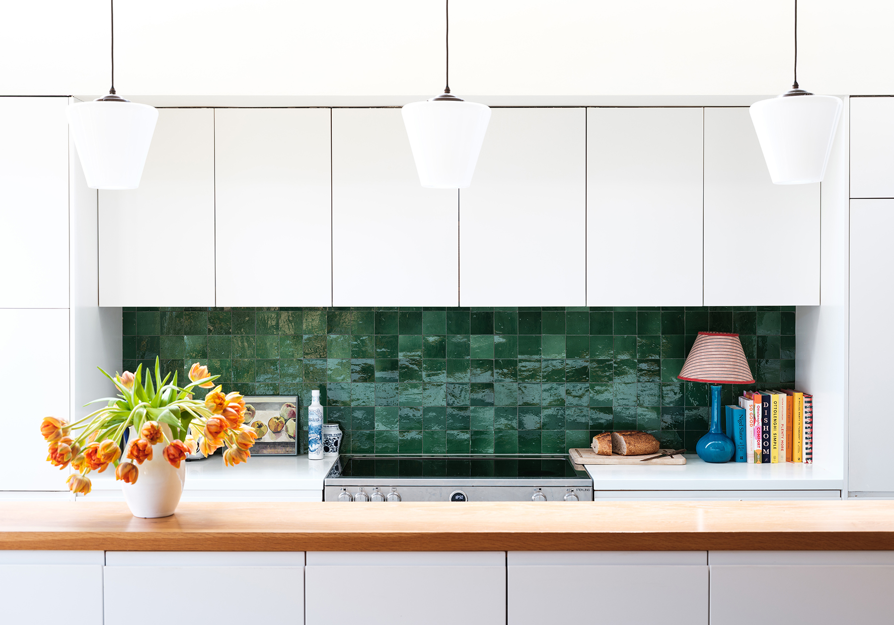 a close up of a kitchen showing white kitchen upper and lower wall cabinets, with light wood worktops, and a backsplash of shiny tiles in differing hues of emerald green, in the foreground a white vase filled with orange tulips sits on top of the worktop