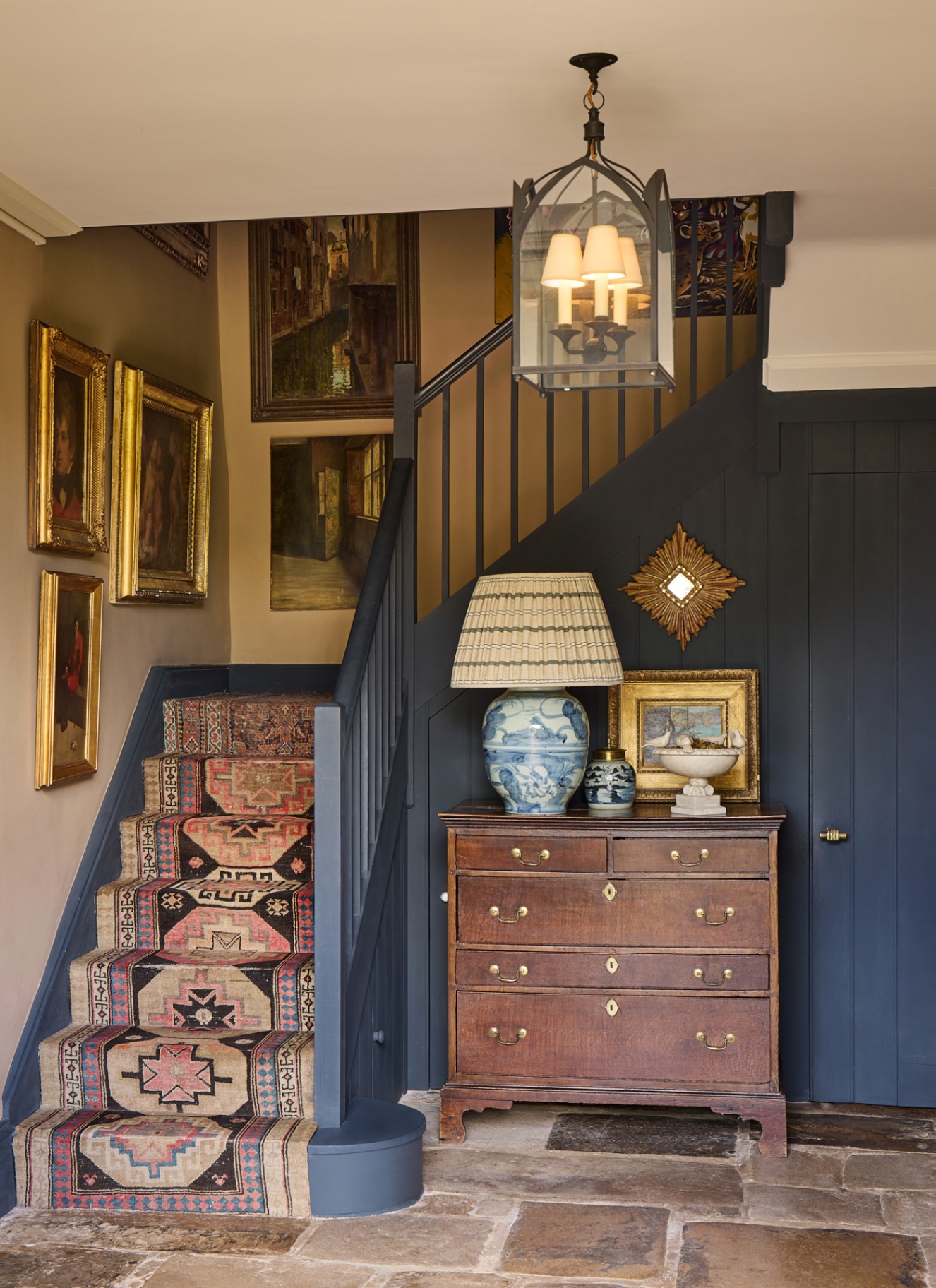 a hallway with blue painted stair banister, vintage patterned stair runner and a vintage chest of draws with a pleated lamp