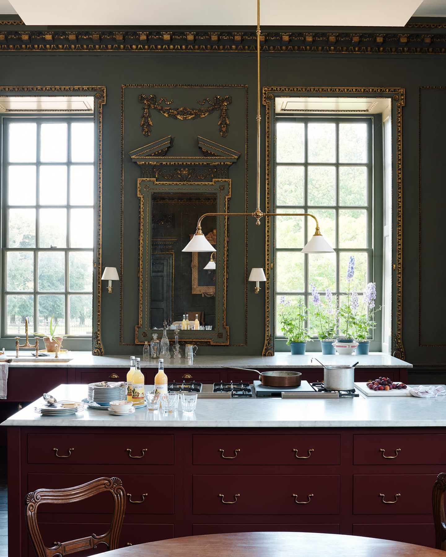 a traditional kitchen with a dark red kitchen island with a large light hanging above it, a wall painted in dark green with gold gilded detailing
