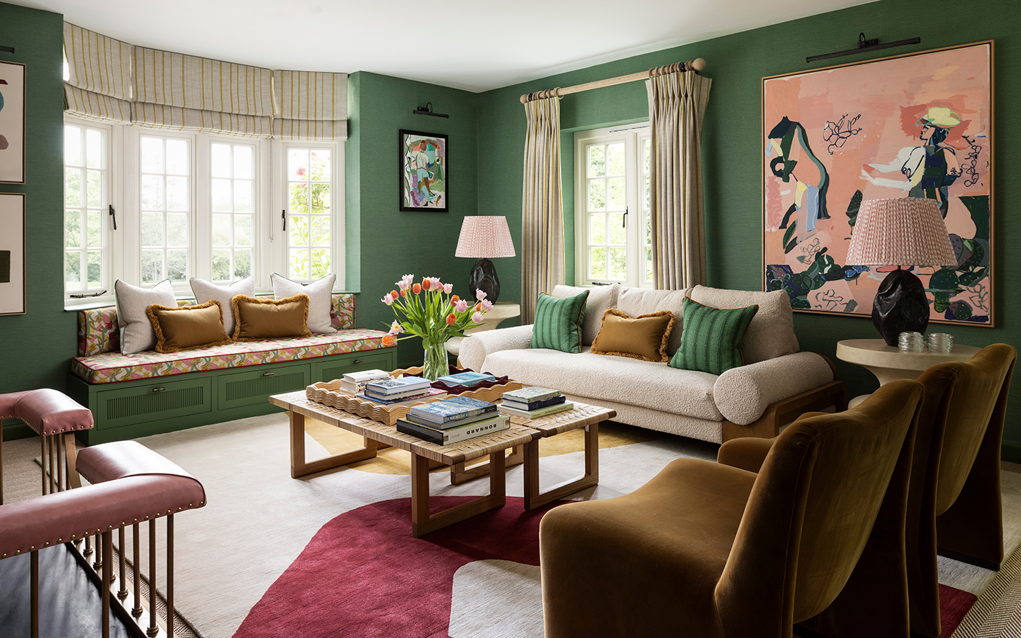 a cosy living room with dark green walls, white boucle sofa, built-in upholstered bench and mustard yellow accent armchairs arranged around a coffee table, with a bold graphic rug underfoot. A large pink abstract painting hangs on the back wall