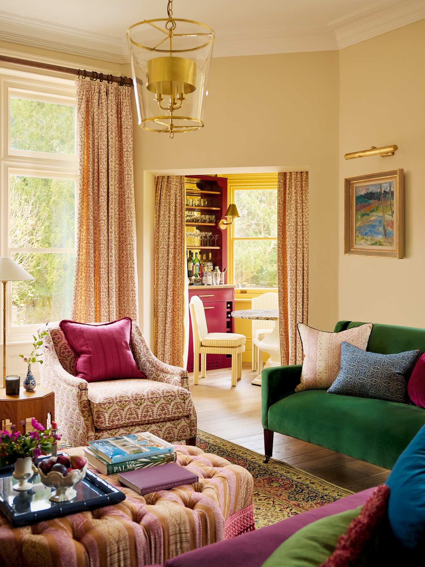 In the foreground is a corner of a living room with two sofas and an armchair around an ottoman,, and adjoining the room in the background is a small octagonal room housing a marble topped table, striped yellow dining chairs and a built-in bar