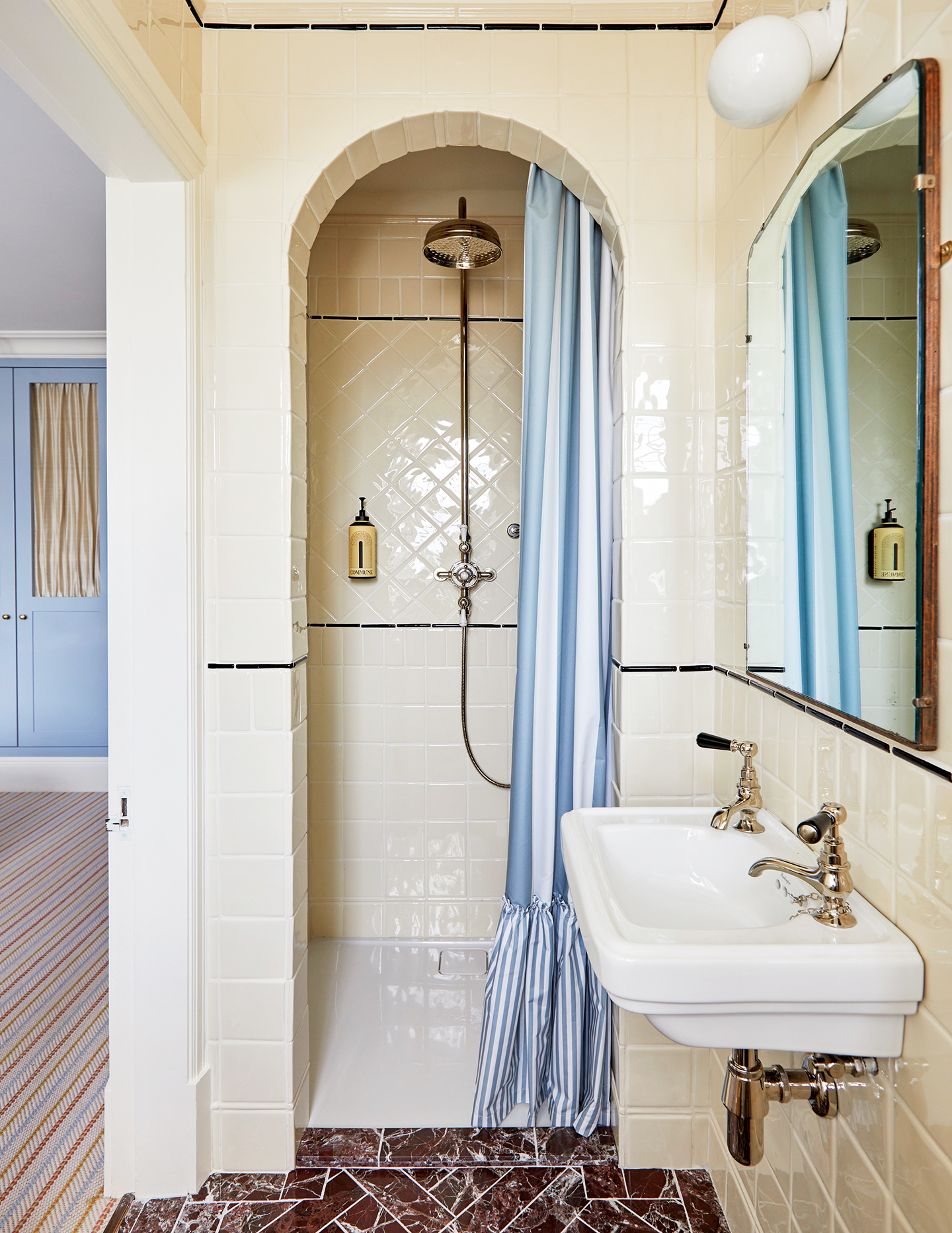 A tiled shower ensuite with an arched shower nook dressed with a blue-and-white striped, frilled shower curtain. A wall-mounted basin sits in the foreground with a mirror and globe wall light above
