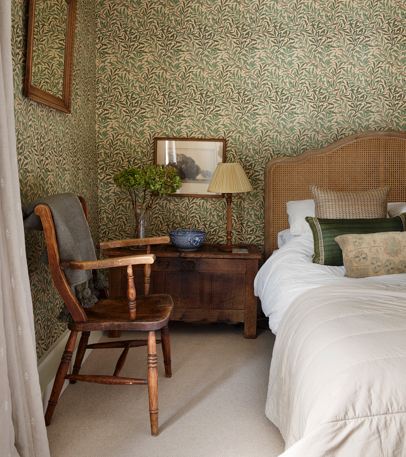 a bedroom with cream and green botanical wallpaper, a wooden low chest of drawers and a wooden armchair next to rattan and wood bedwith white bedding and beige and green cushions