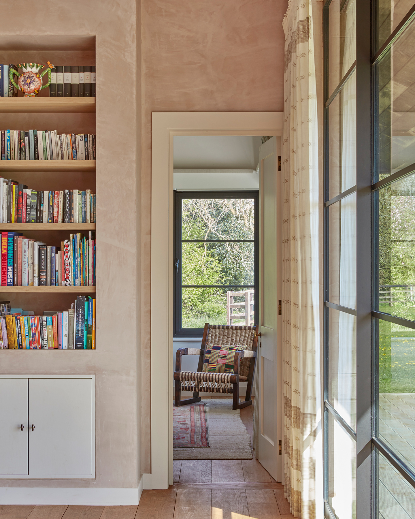 A view through a door to a room with a woven wicker chair topped with a cushion and a vintage rug on the wood floors. In the foreground, shelving built in to raw plaster walls holds books and glazing on the right is dressed with a neutral curtain