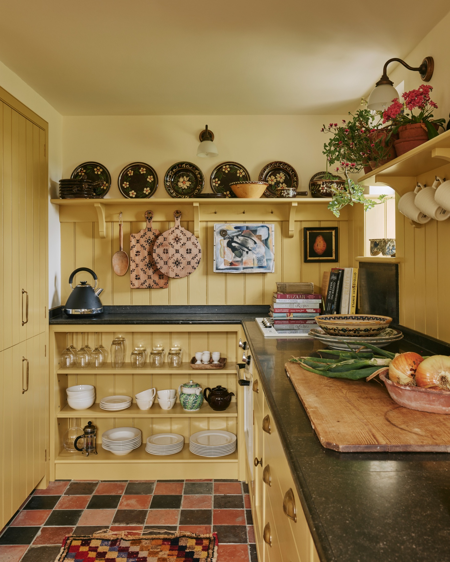 a country kitchen with lemon yellow panelled walls, orange and black teracotta floor tiles and open shelving with decorative plates