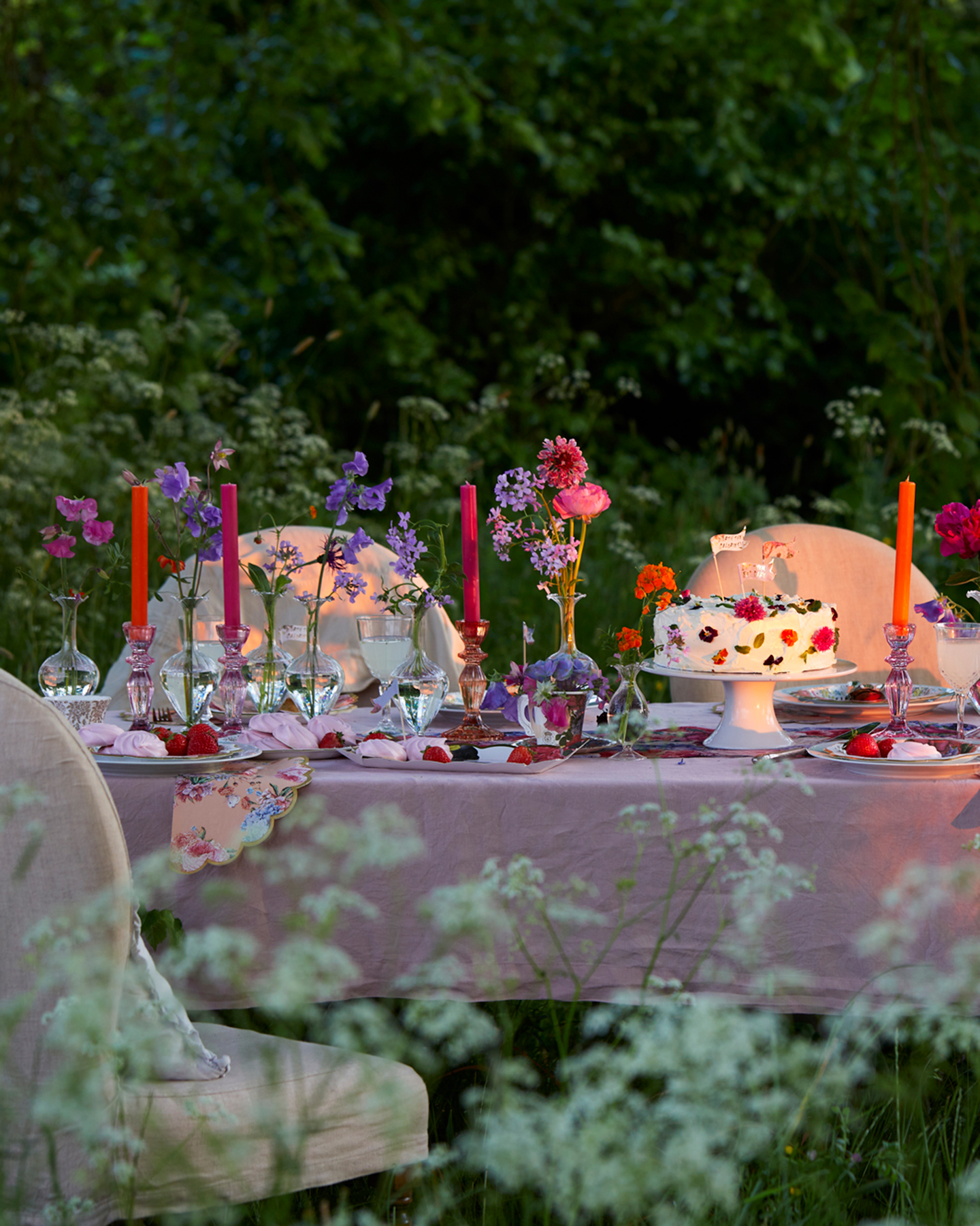 an outdoor table with a pink table cloth and pale pink upholstered chairs. A floral cake on a cake stand on top of the table with small vases of flowers and bright orange and pink dinner candles in glass holders