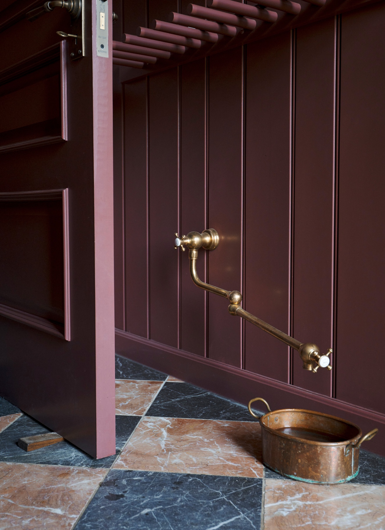 a floor level long brass tap feeding into a brass dog bowl against a dark red wall on a checkered tile floor
