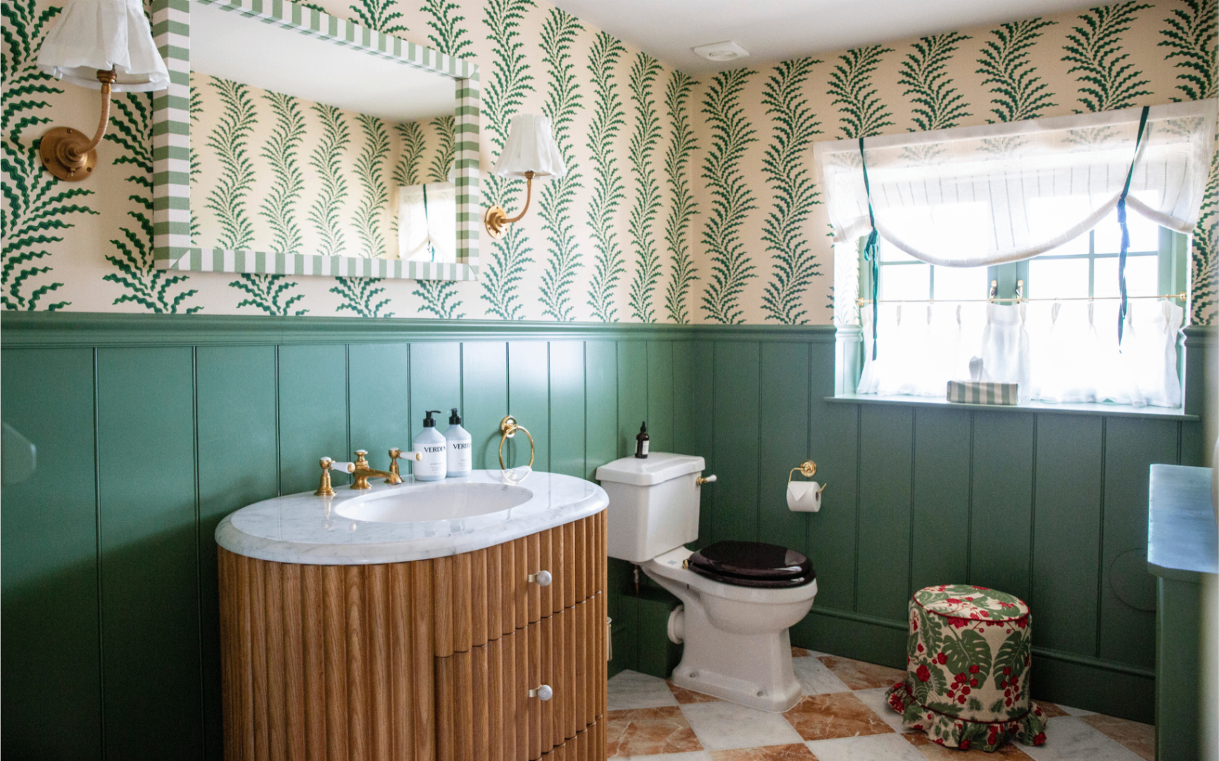 a downstairs toilet with half teal panelled walls, half cream and green vine wallpaper, a fluted wooden vanity sink with a white and green striped mirror above it with two pleated shade wall lights