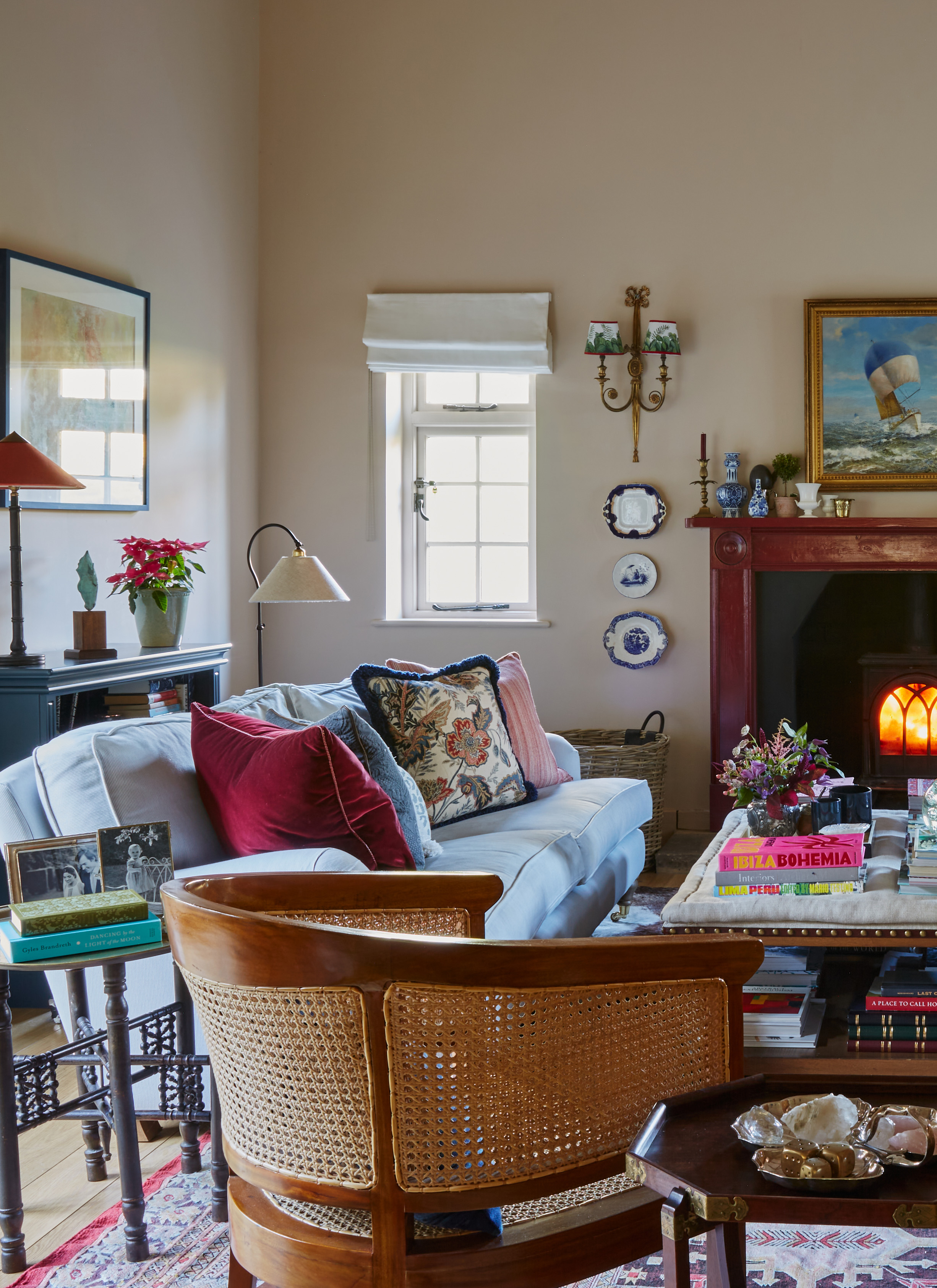 A neutral-toned sitting room with a blue sofa, a cane back armchair, wall sconces, wall plates, and a dark wooden fireplace mantle with a burning fire